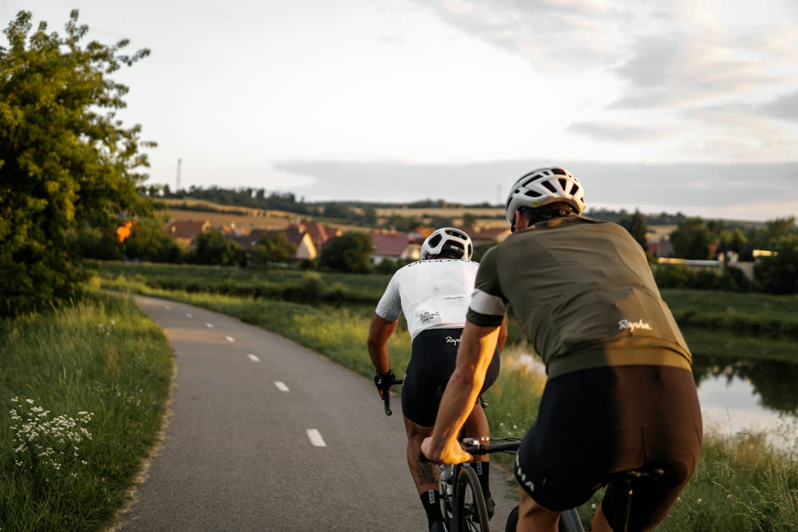 Two road cyclists seen from behind, riding a winding country lane at golden hour toward a village in the valley below.