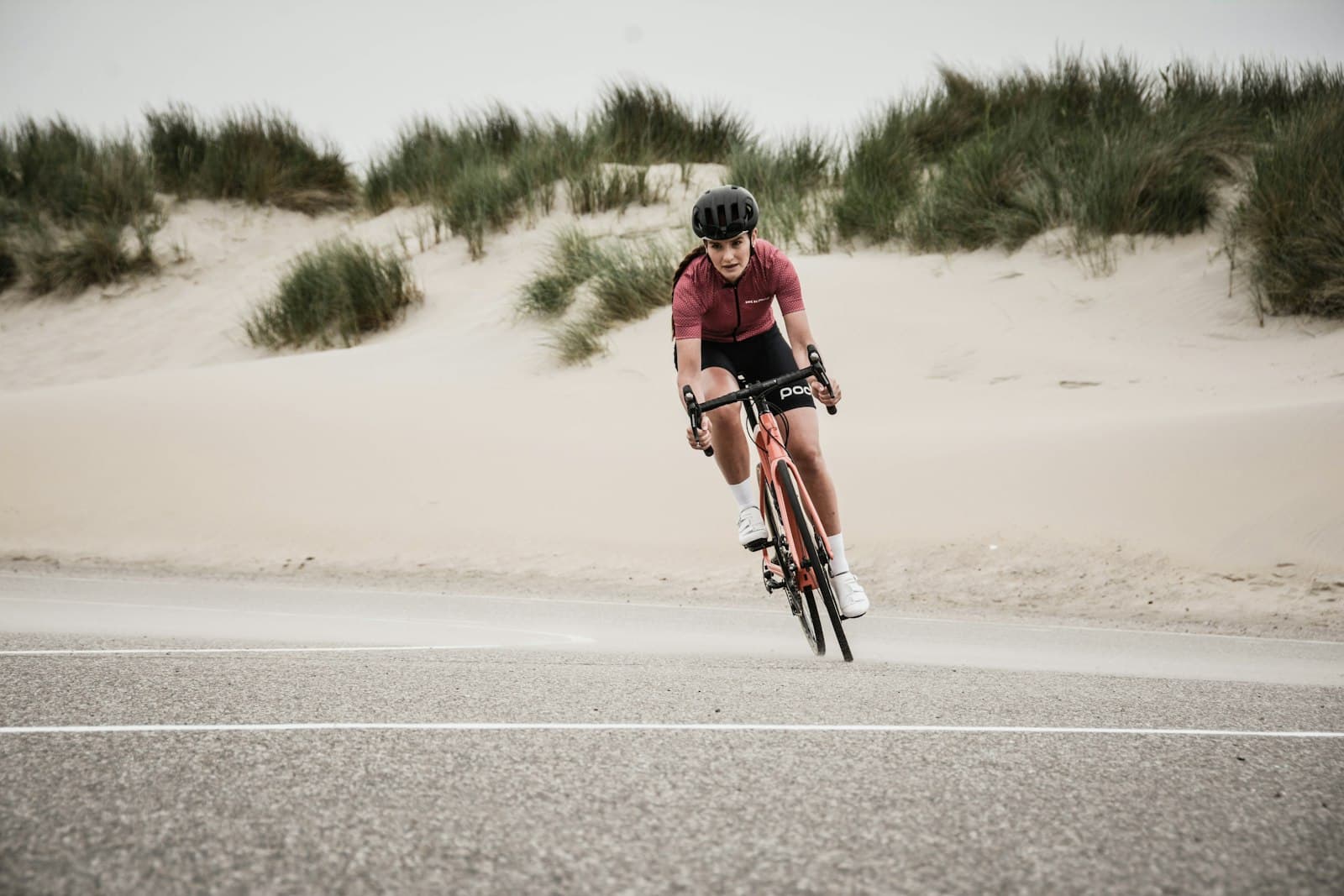 A road cyclist in a maroon jersey riding solo on an open coastal road, sand dunes behind her under an overcast sky.